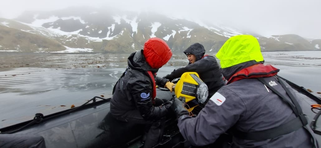 Three people in a zodiac, wearing wet-weather gear, lifting a buoy into the water.