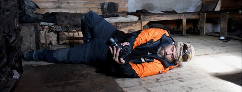 A man laying on his side, taking a photograph. Antarctic Heritage Trust.
