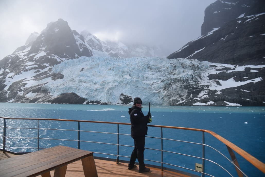 A woman on a boat holding a 360 degree camera with a glacier in the background.