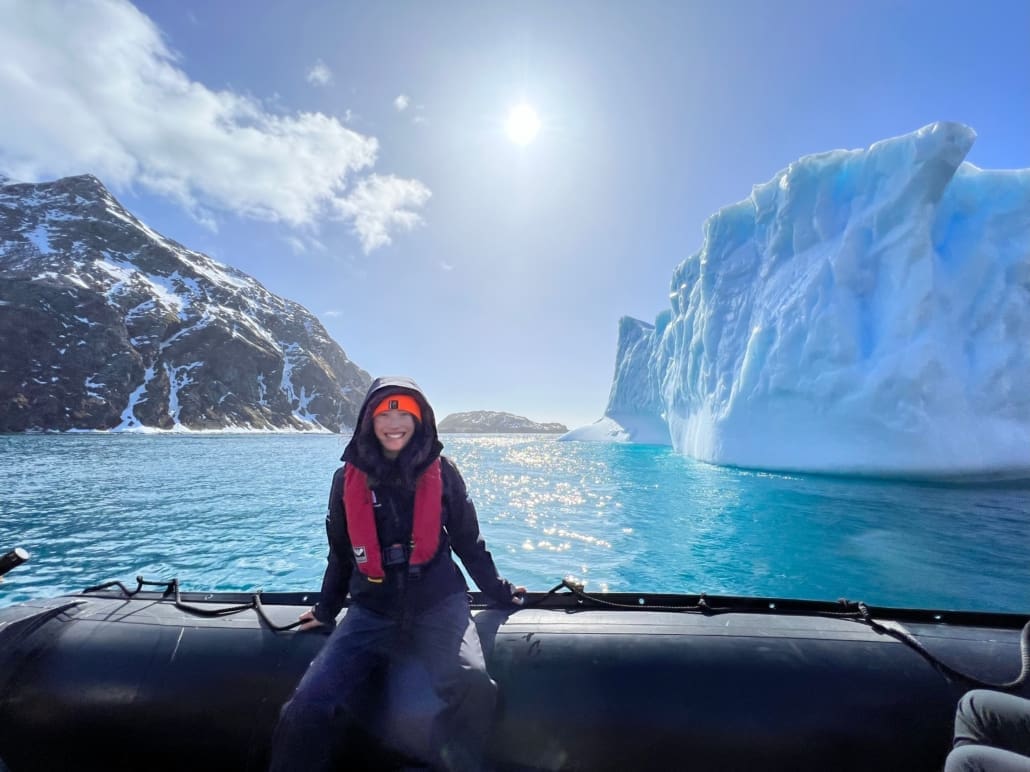 A woman sits on the edge of a boat as it floats in front of large icebergs and snowy mountains.