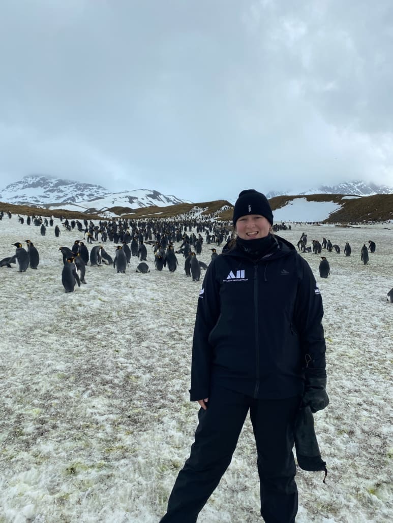 A woman stands in an icey landscape with mountains and thousands of penguins behind her.