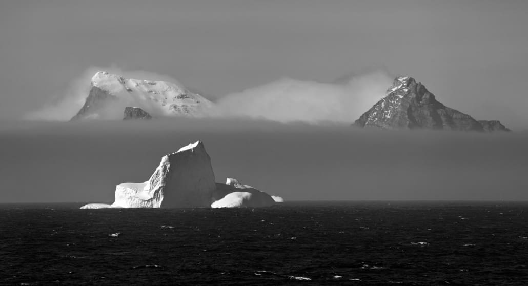 An iceberg in water, with two mountain peaks appearing above low cloud in the background.