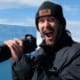 A man with an excited expression holds a camera, with giant icebergs in the background.