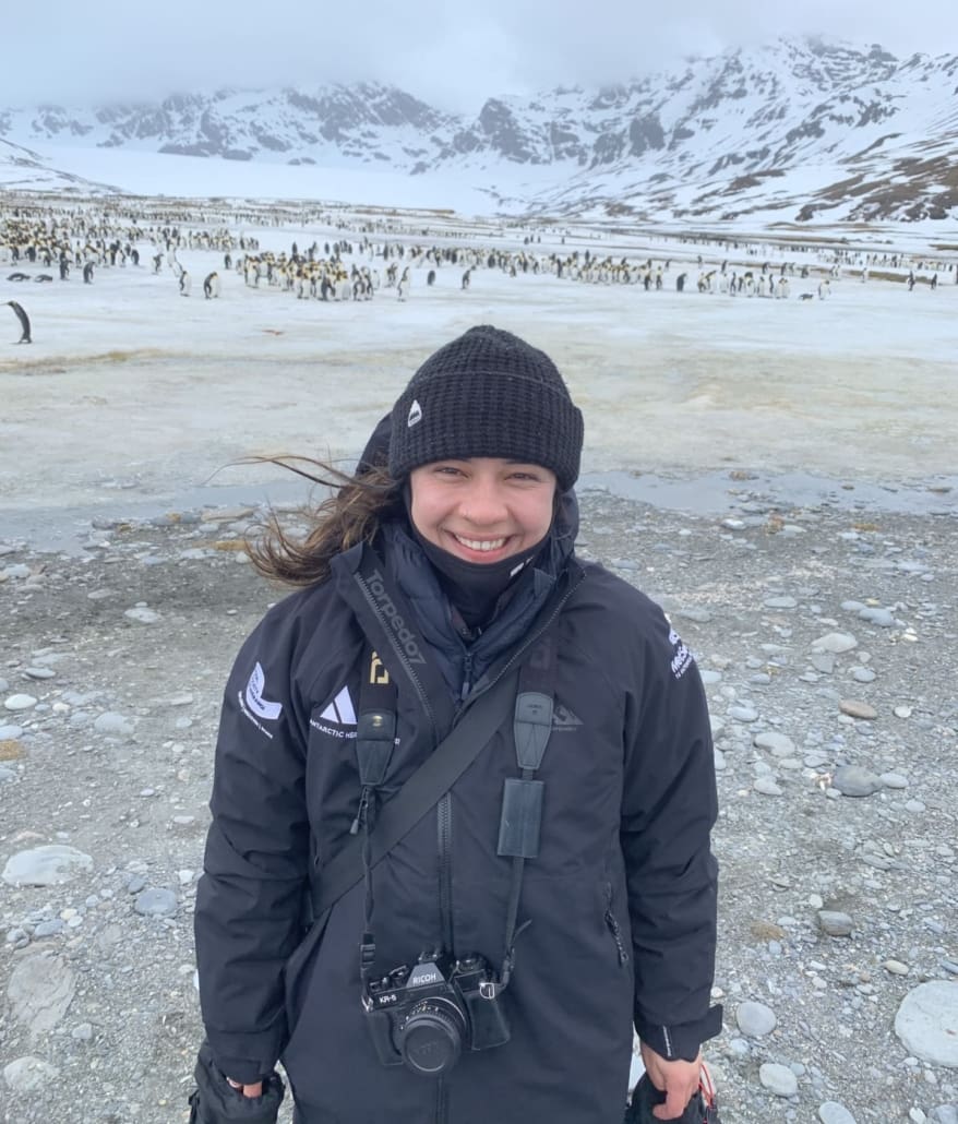 A woman smiles with snowy mountains and thousands of penguins in the distance behind her.