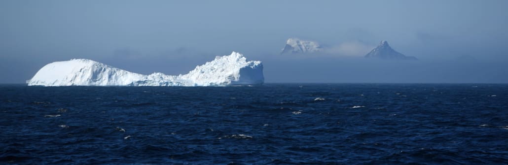 A large iceberg in ocean waters with mountains peaks shrouded in cloud in the background.